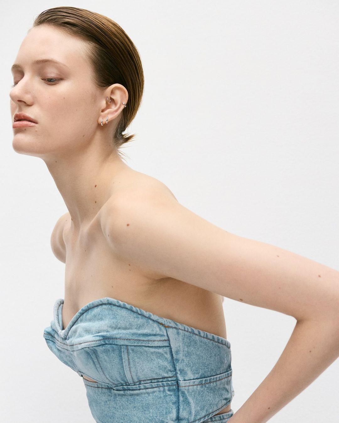 Profile portrait of a young adult female with slicked-back hair wearing a denim top against a white background in a photo studio.
