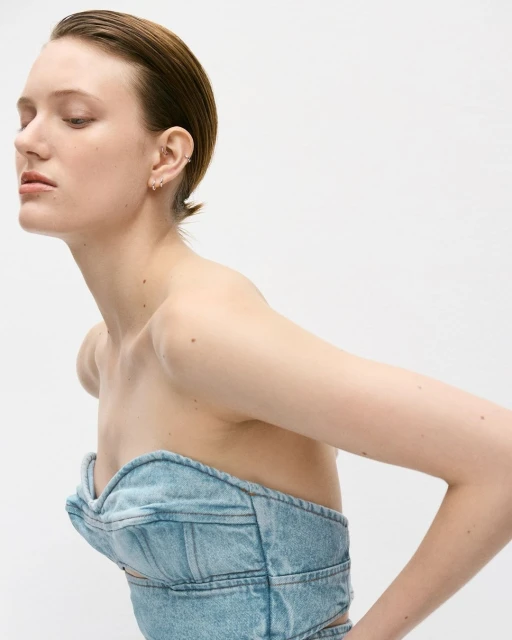 Profile portrait of a young adult female with slicked-back hair wearing a denim top against a white background in a photo studio.