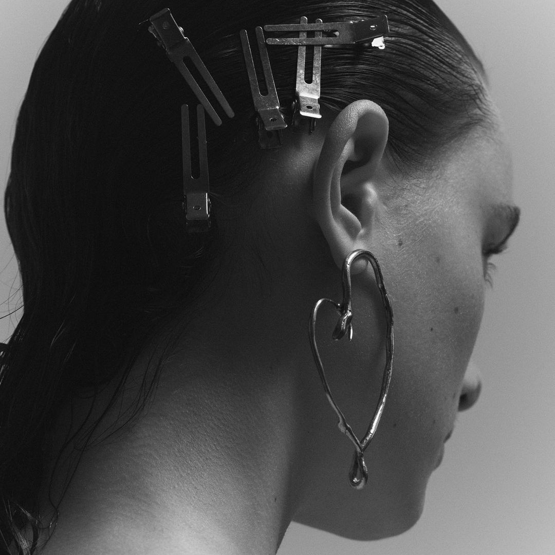 Close-up black and white studio shot of a woman with hair clips and a heart-shaped earring, minimalist aesthetic, fashion-forward jewelry.