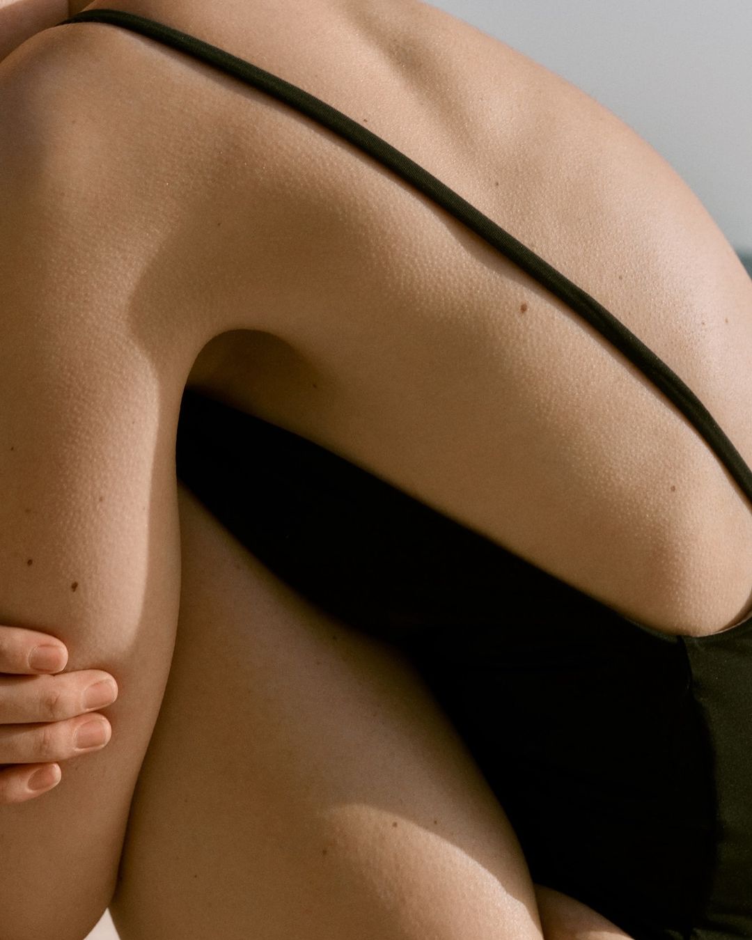 Close-up of a tanned adult female's back and arm. She is wearing a black swimsuit and sitting in a sunny, outdoor environment.