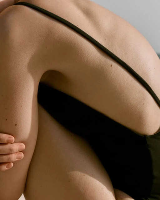 Close-up of a tanned adult female's back and arm. She is wearing a black swimsuit and sitting in a sunny, outdoor environment.
