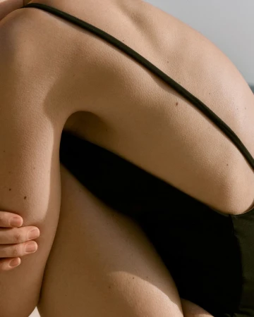 Close-up of a tanned adult female's back and arm. She is wearing a black swimsuit and sitting in a sunny, outdoor environment.