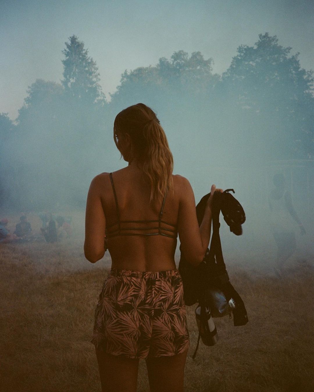 A woman from the back in a field near a forest holds a black backpack, creating a natural and serene scene.