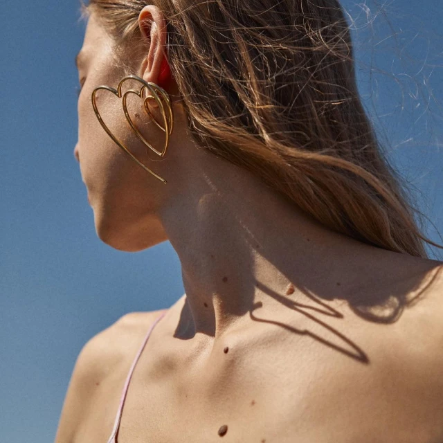 Close-up of a woman wearing large gold heart-shaped earrings against a bright blue sky, showcasing fashion jewelry.