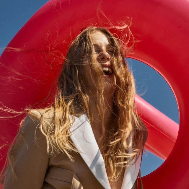 Smiling woman with long hair, wearing a blazer, is seen from a low angle against a vivid red inflatable and a clear blue sky.