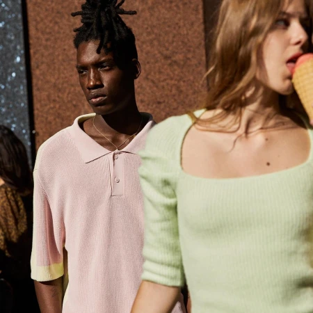 Two people standing outdoors; one wearing a light pink polo shirt and the other eating an ice cream cone in a green knit top.