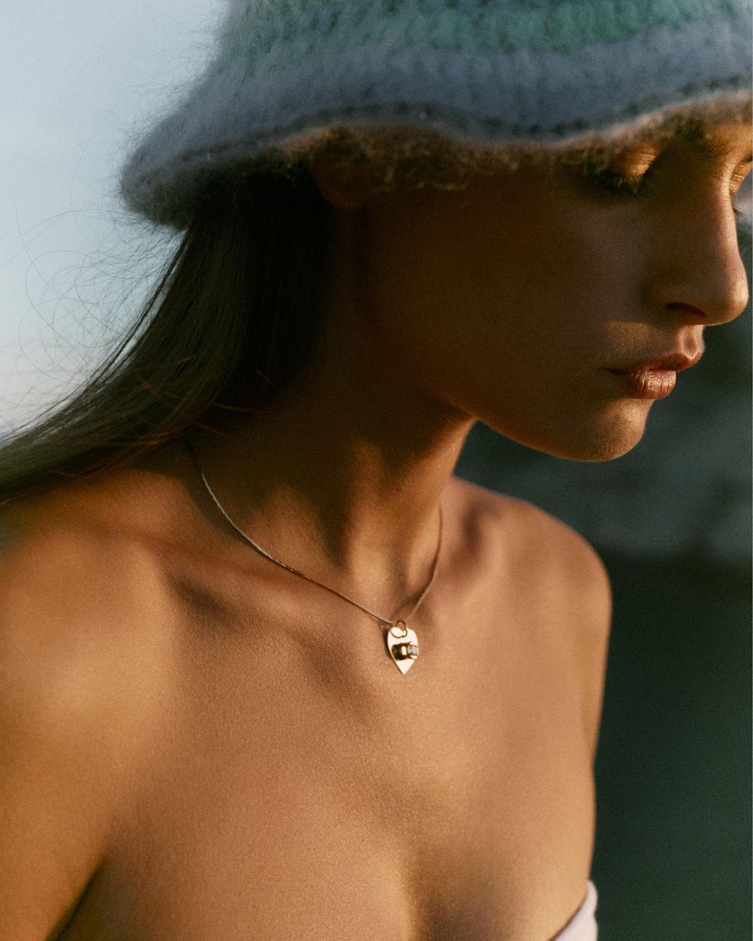Portrait of woman wearing light-colored bucket hat and gold pendant necklace, with soft lighting and shallow depth of field.