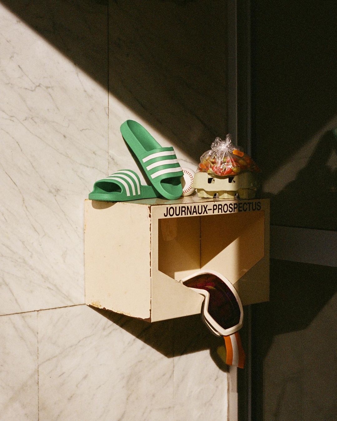 Still life composition featuring green Adidas slides, a baseball, an egg carton with wrapped fruit, and ski goggles, displayed on a wall-mounted box.