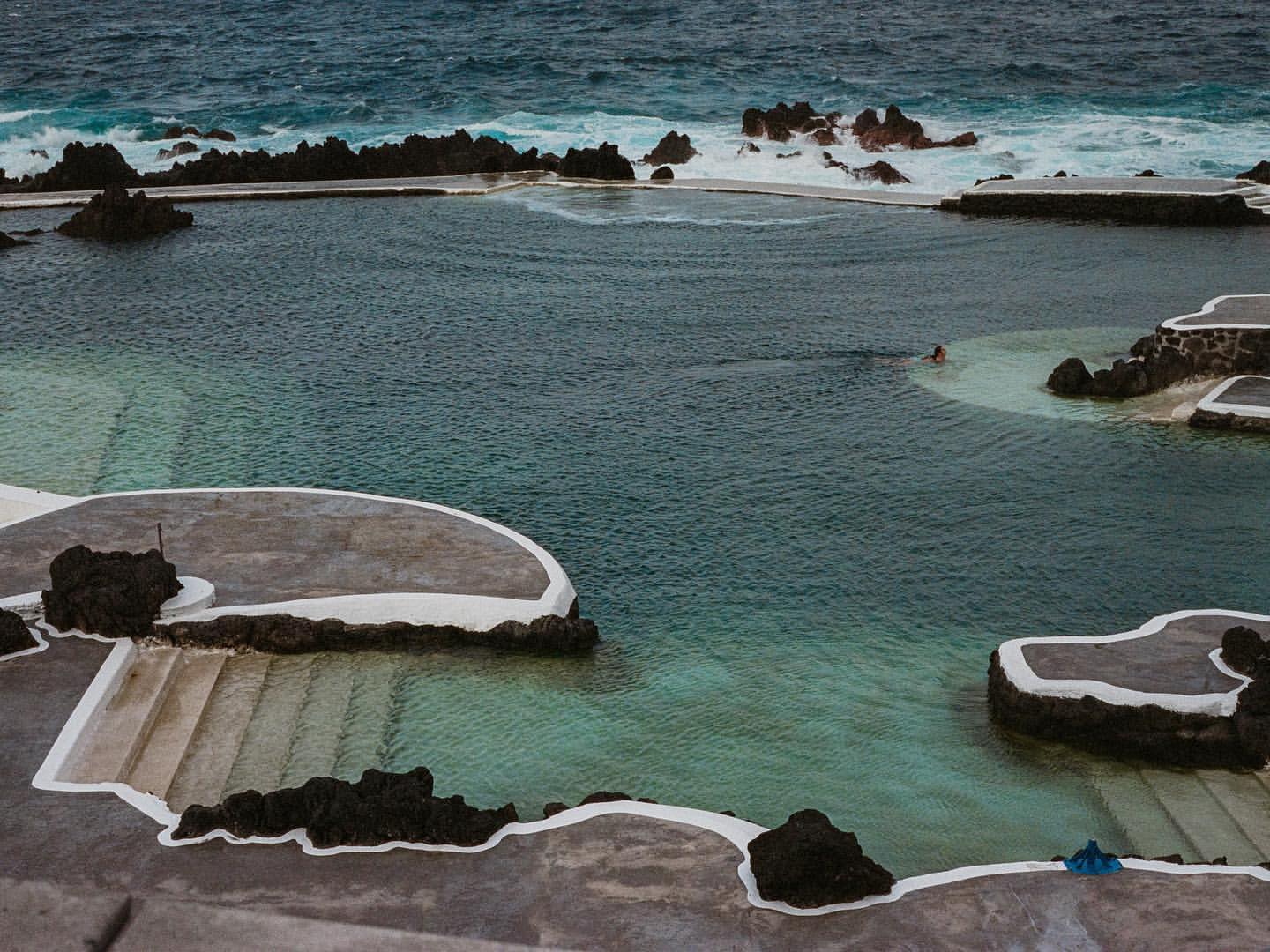 High-angle shot of natural rock pools filled with ocean water, featuring volcanic rock formations and ocean waves in the background.
