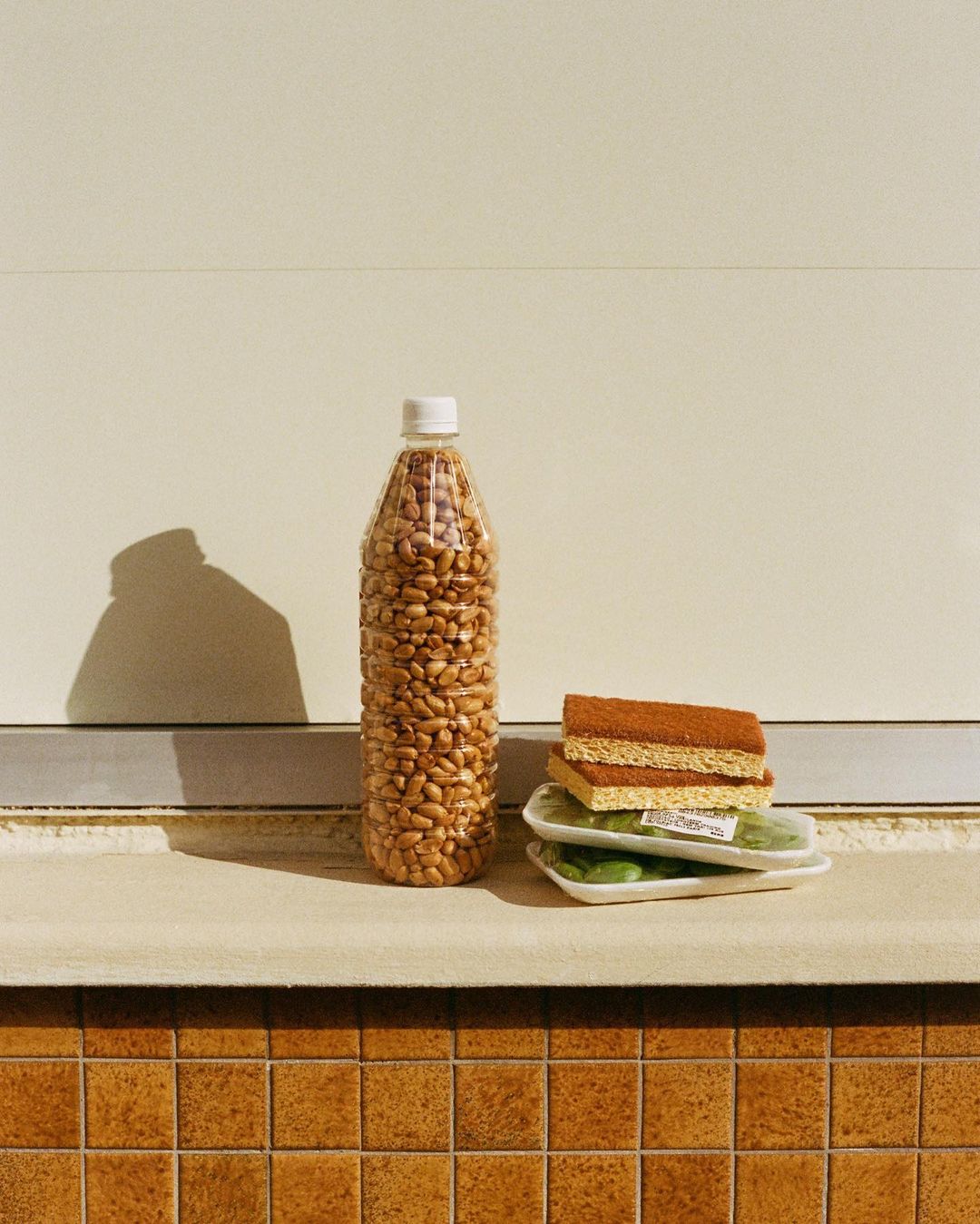 Still life of a plastic bottle filled with beans, stacked cleaning sponges, and produce trays on a ledge against a tile wall backdrop.