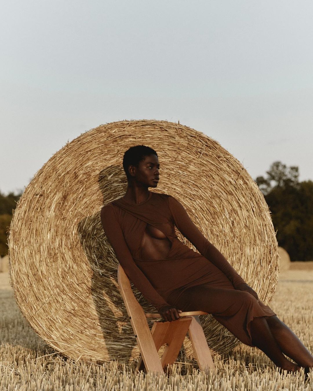 An African American woman in a brown dress poses seated in front of a hay bale in a wheat field on a sunny day, fashion shot.