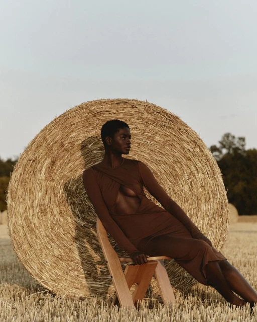 An African American woman in a brown dress poses seated in front of a hay bale in a wheat field on a sunny day, fashion shot.