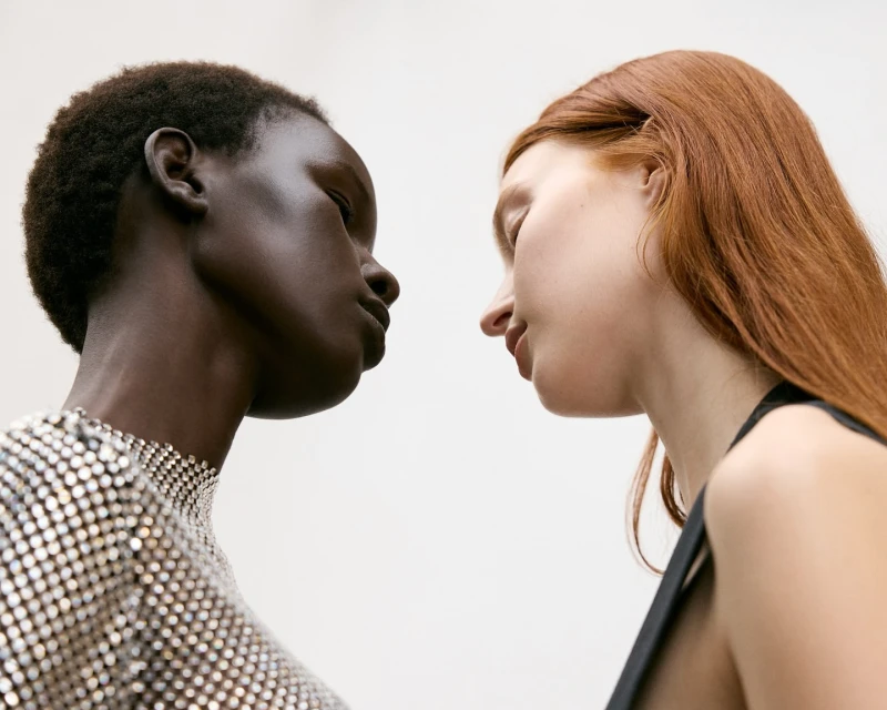 Two diverse women facing each other in a close-up portrait. The woman on the left has dark skin, while the one on the right has fair skin.