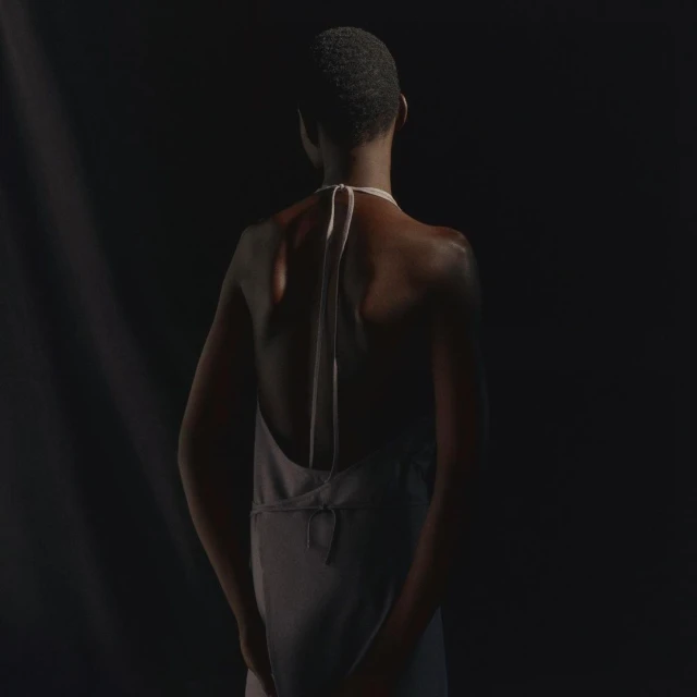 Back view of a slender adult fashion model in a light grey halter dress, standing in a dark photo studio with soft directional lighting.