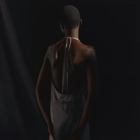 Back view of a slender adult fashion model in a light grey halter dress, standing in a dark photo studio with soft directional lighting.