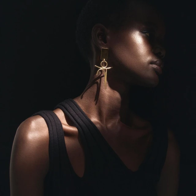 Close-up studio shot of a Black woman with a gold earring in a black sleeveless dress against a dark background. Dramatic lighting.