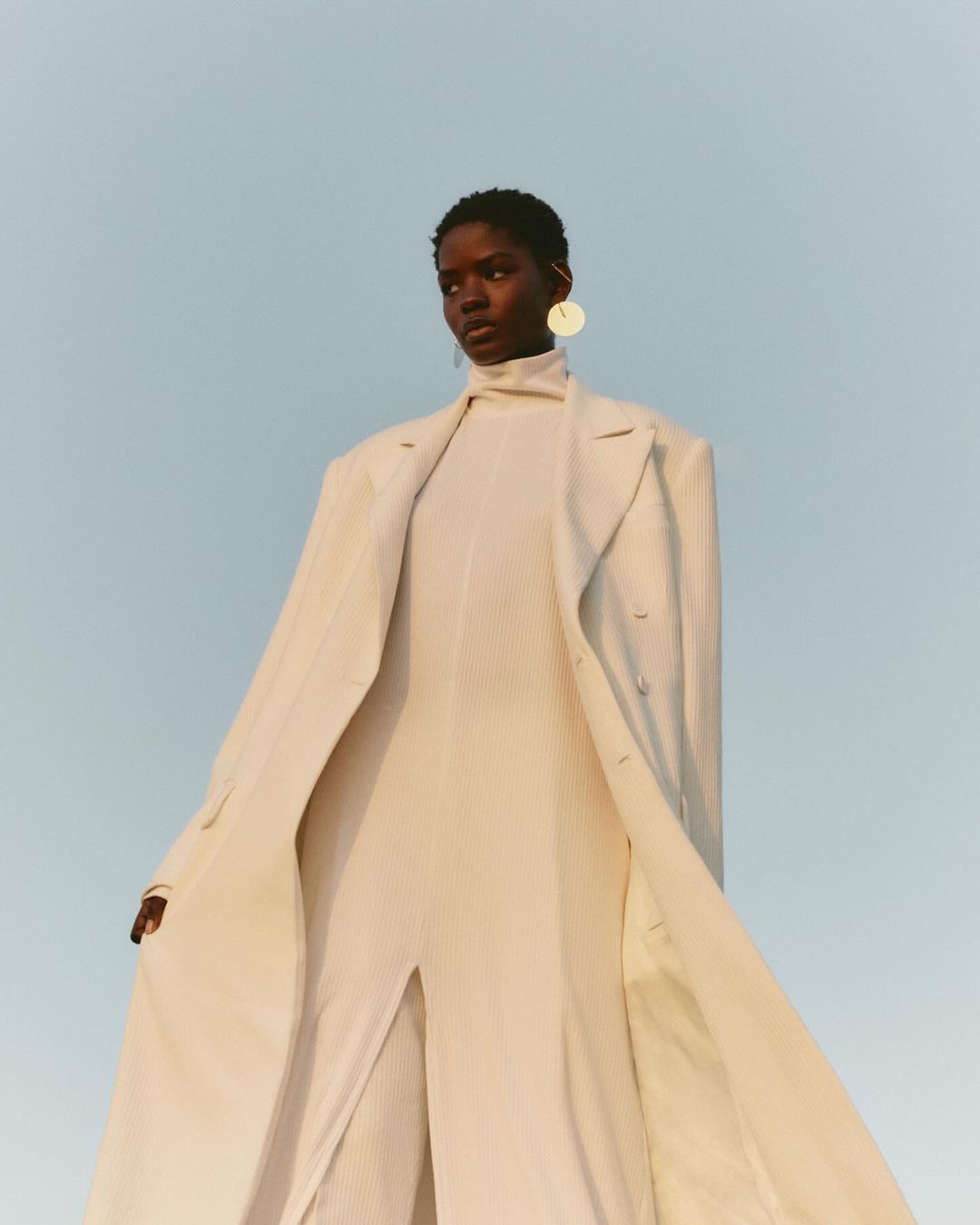 Elegant woman in a full-length coat and jumpsuit with a neutral backdrop, wearing statement earrings for a fashion-forward aesthetic.