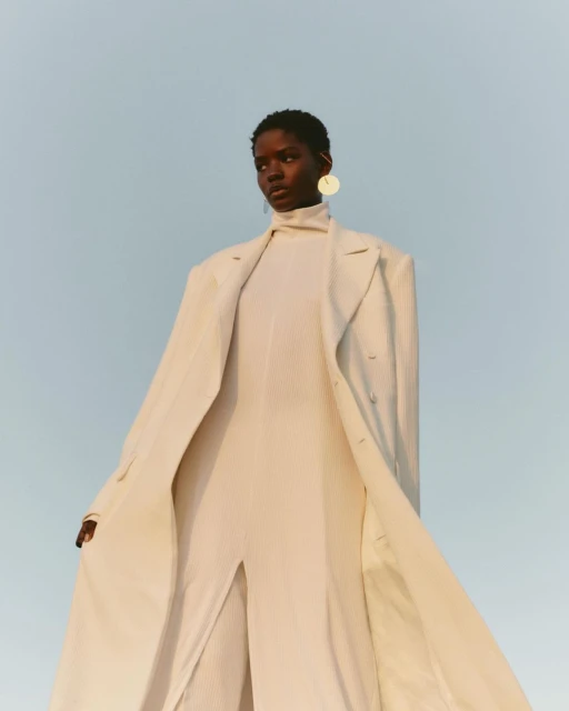Elegant woman in a full-length coat and jumpsuit with a neutral backdrop, wearing statement earrings for a fashion-forward aesthetic.
