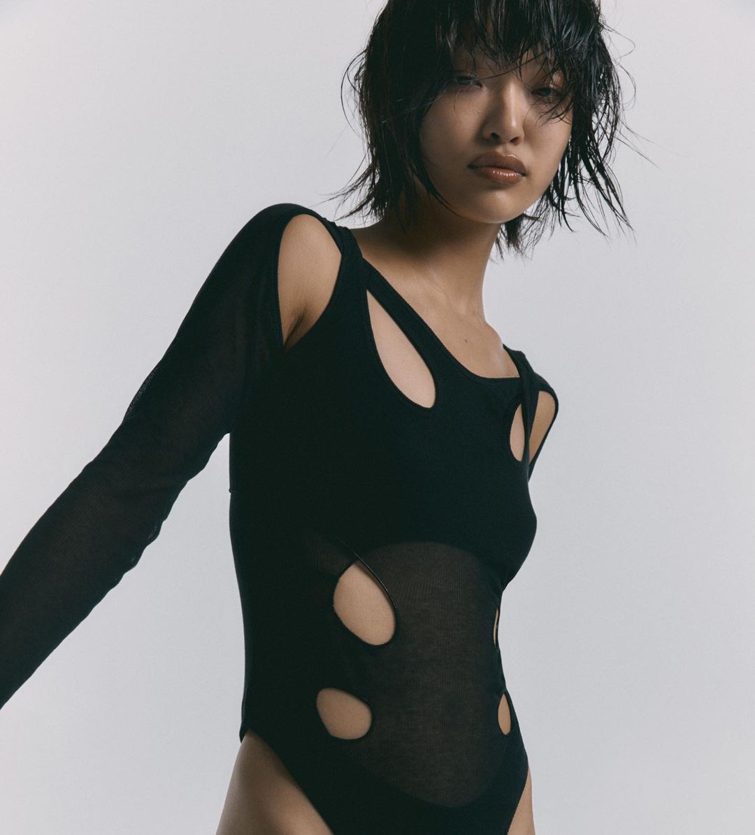 Young woman with wet, dark hair in a black cut-out leotard against a white background, posing in a studio setting. Fashion shot.
