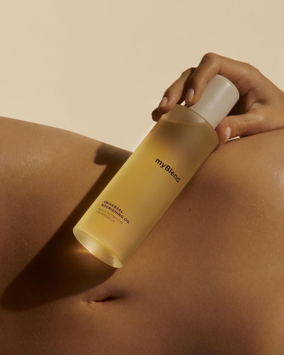 Close-up studio shot of a woman's torso holding a bottle of clear body oil against a beige background. Bodycare and skincare concept.