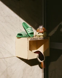 Green slider sandals and ski goggles arranged on top of a cream wall-mounted newspaper box in Paris.