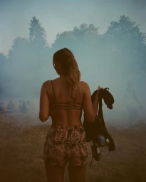 Woman viewed from behind wearing a patterned crop top and shorts holding a jacket in a hazy park setting.