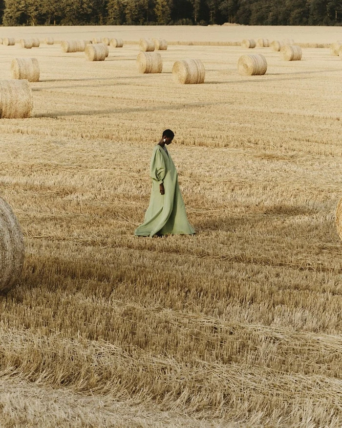 Model in long sage green dress walking through a harvested field dotted with round hay bales under natural daylight.