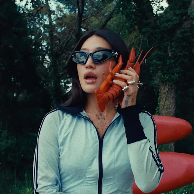Woman in sunglasses holds a lobster to her ear as if it's a phone, with a green wooded area and a red object in the background.