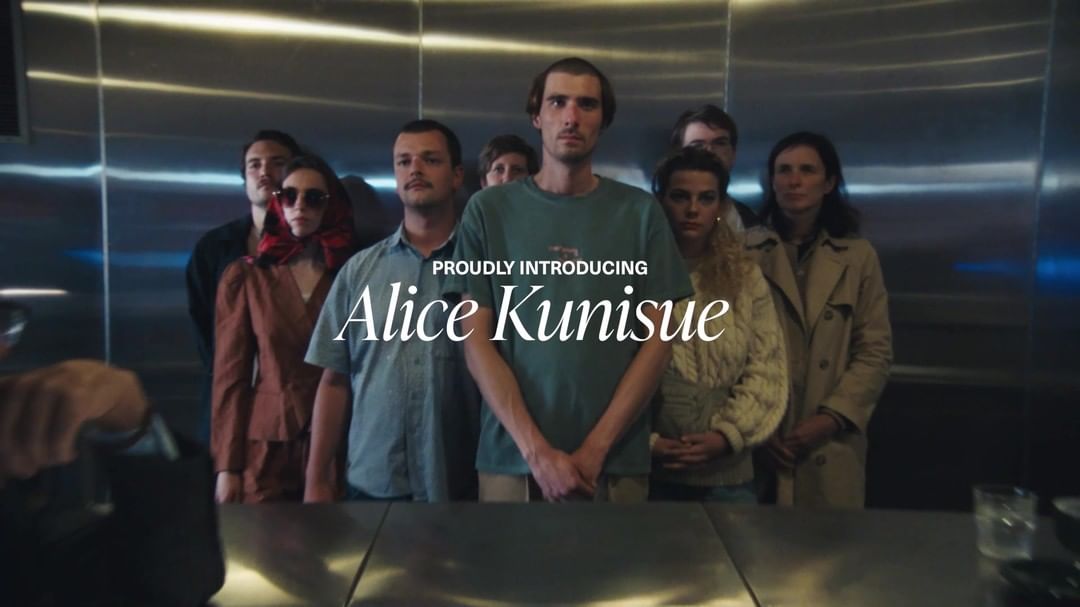 Group portrait of eight adults in casual clothing, posing in front of a metallic elevator with text overlay 'Proudly Introducing Alice Kunisue'.