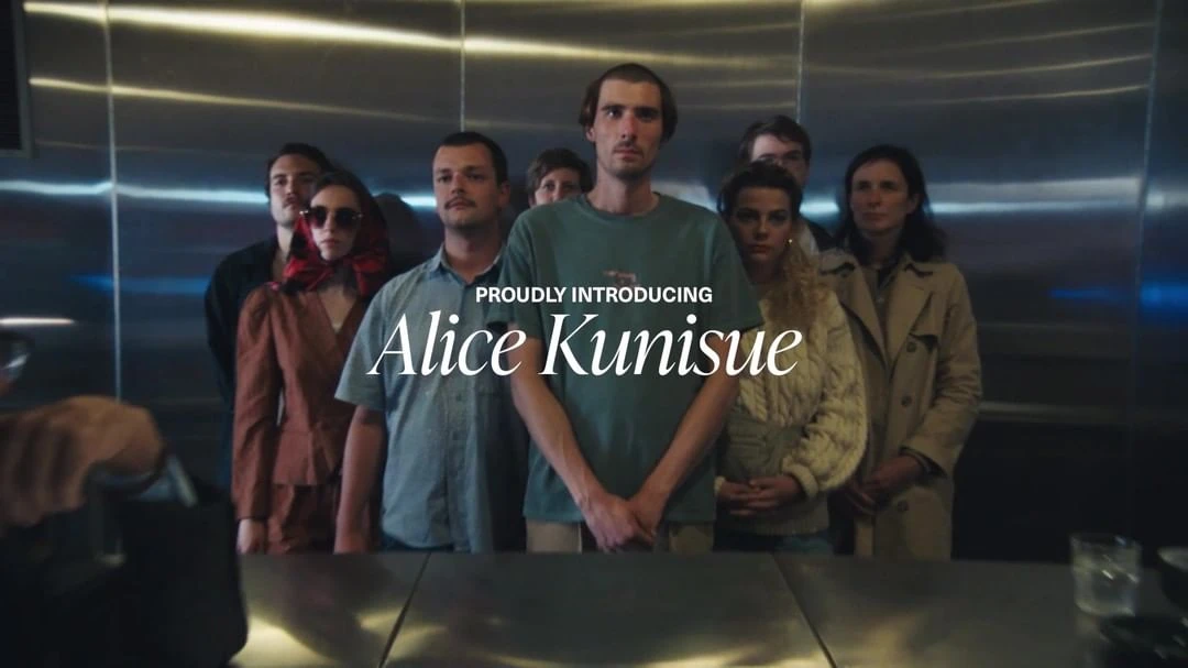 Group portrait of eight adults in casual clothing, posing in front of a metallic elevator with text overlay 'Proudly Introducing Alice Kunisue'.