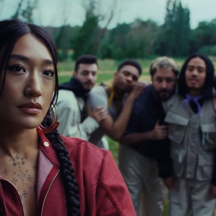 Portrait of an Asian woman in a red jacket with braided hair standing in a field, with four blurred men standing in the background.