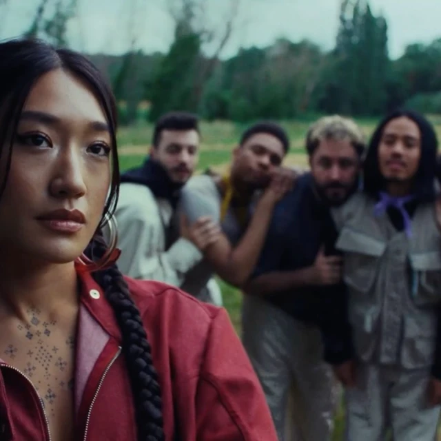 Portrait of an Asian woman in a red jacket with braided hair standing in a field, with four blurred men standing in the background.