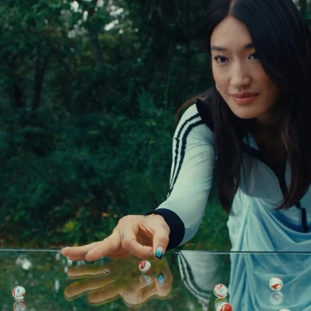 Woman in track suit plays with marbles on glass table in outdoor scene with lush green foliage, showcasing leisure and reflection.