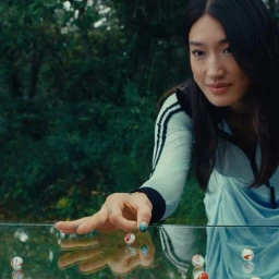 Woman in blue athletic jacket reaching over a reflective glass table containing several small marbles, with green foliage in the background.