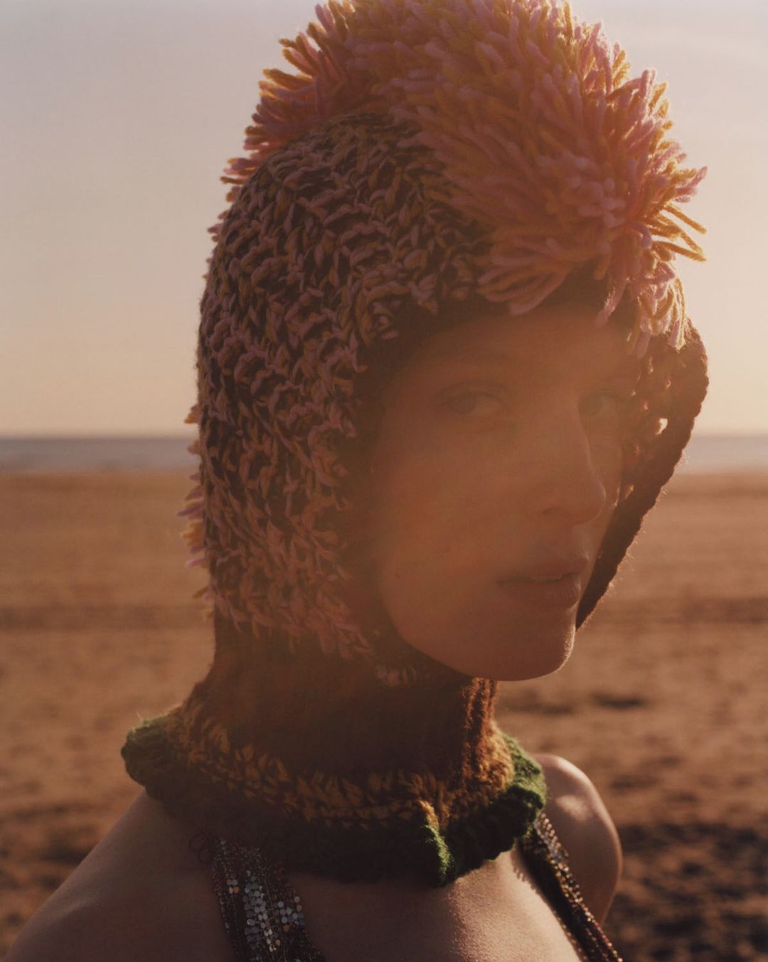 Close-up of a woman in a textured knit hat with spikey embellishments, standing on a sandy beach under an evening sky with soft sunlight.