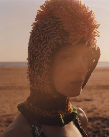 Close-up of a woman in a textured knit hat with spikey embellishments, standing on a sandy beach under an evening sky with soft sunlight.