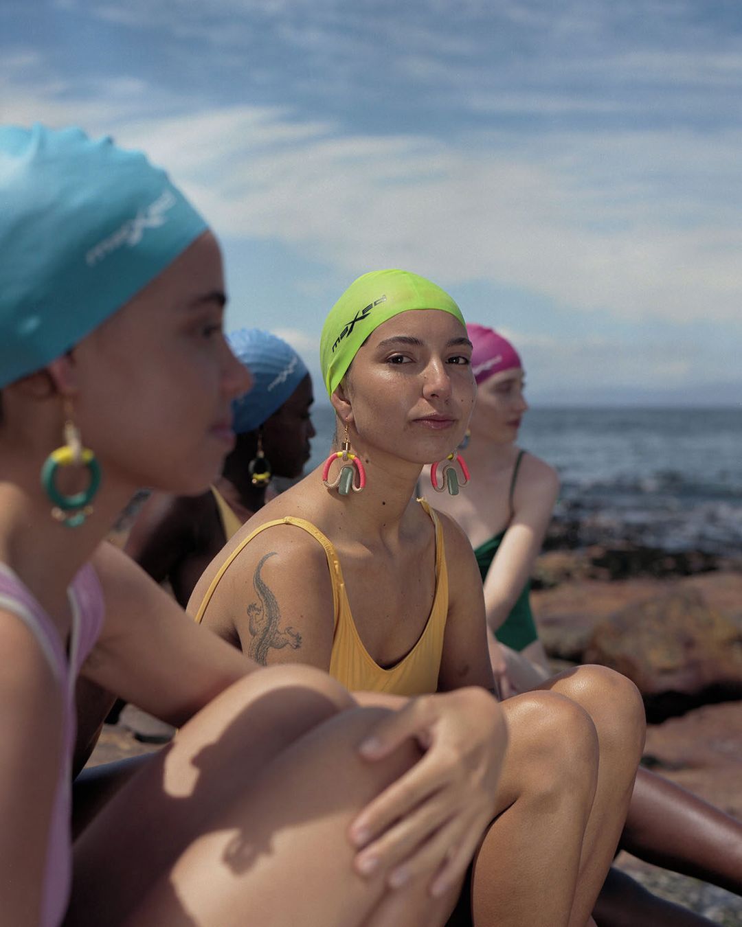 Several women wearing swimsuits and swim caps sit together on rocks near a beach under a cloudy sky. Earrings are visible. 