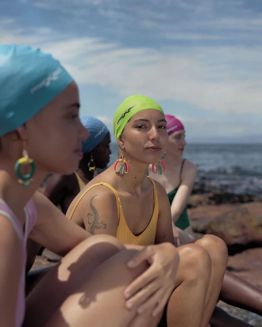Several women wearing swimsuits and swim caps sit together on rocks near a beach under a cloudy sky. Earrings are visible. 
