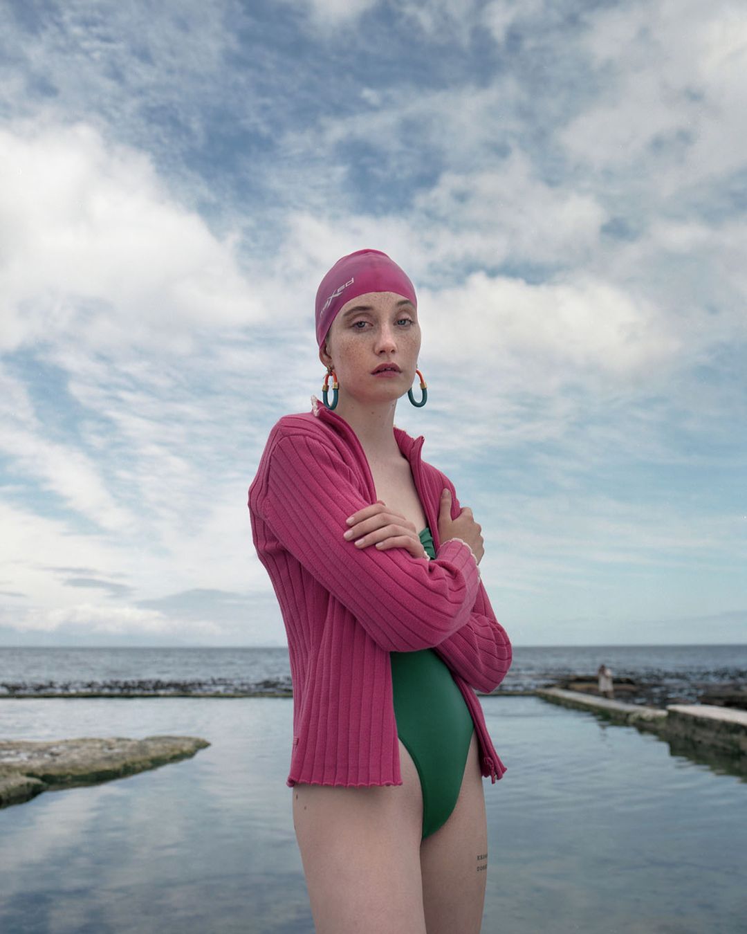 A young adult female wearing a pink swim cap and cardigan stands with crossed arms by a natural pool on a cloudy day, portrait shot.