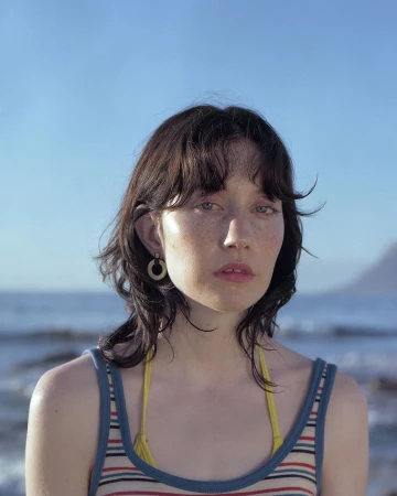 Portrait of a young woman with dark hair and freckles wearing a striped tank top outdoors near the ocean.