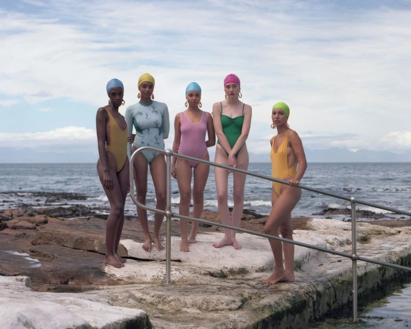 Group of five women in swimsuits and colorful swim caps stand on a stone platform near the ocean under a cloudy sky.