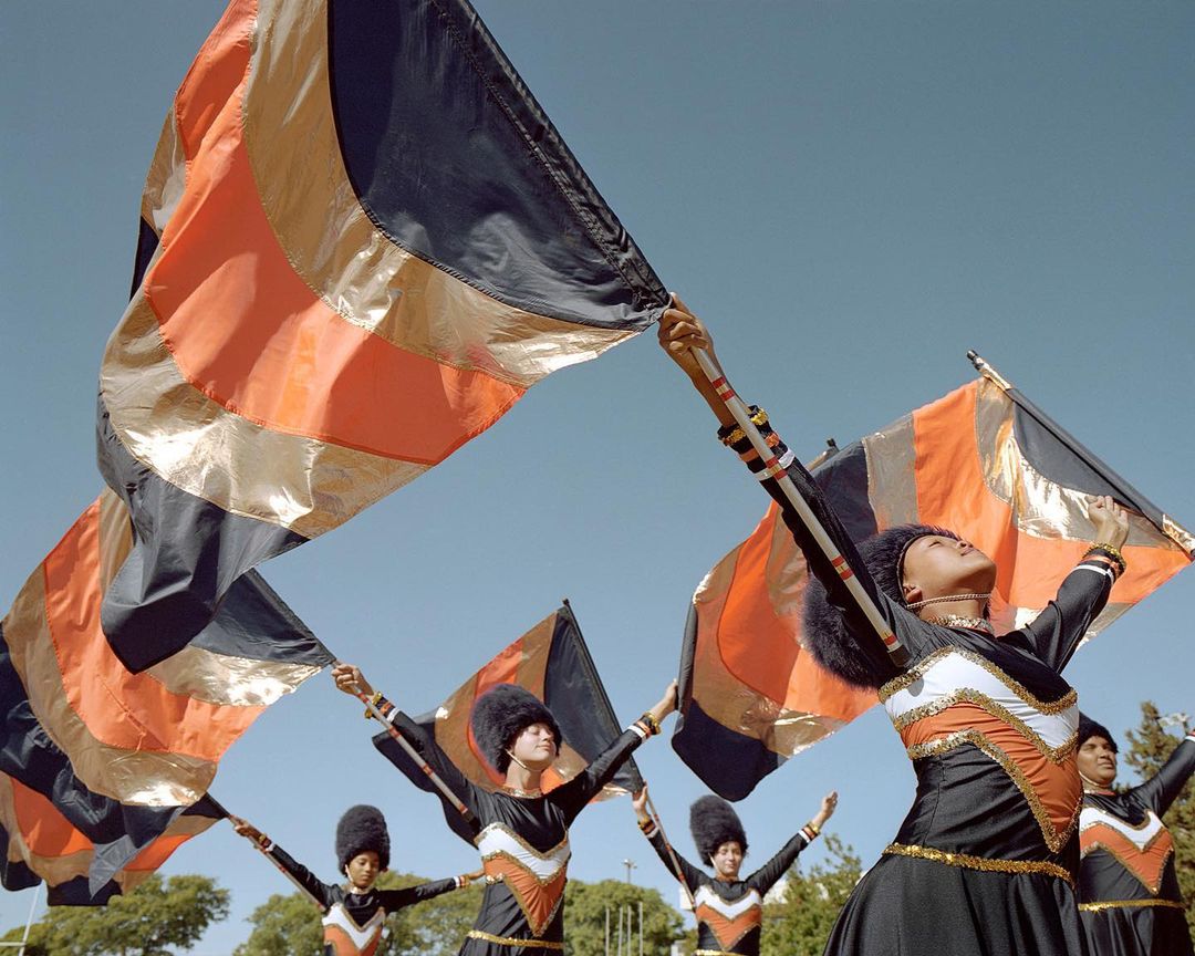 Group of color guard performers waving flags in unison with black, orange, and gold colors against a clear blue sky on a sunny day.