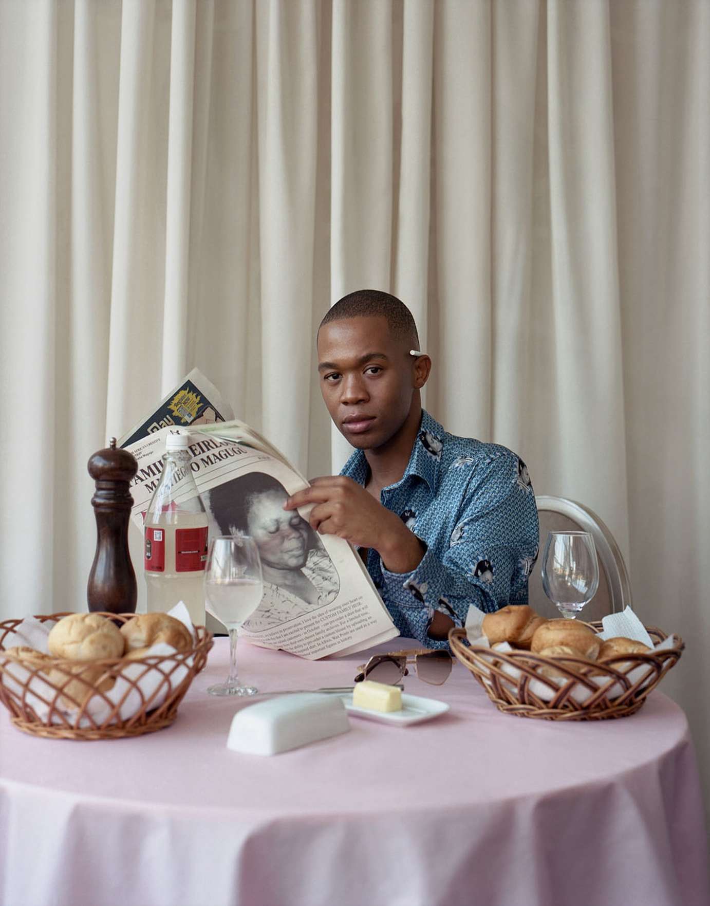 Person at a table reading a newspaper with bread baskets, drinks, butter, and a pepper grinder, sitting in front of cream drapes.