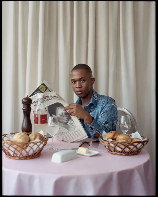Adult male reading newspaper at table set with bread, wine, butter, and water, in front of a cream-colored curtain backdrop. Indoor setting.