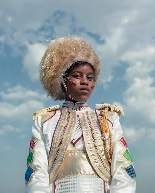 Young person in an ornate white costume, decorative hat, and facial markings poses against a partly cloudy sky in a medium shot.