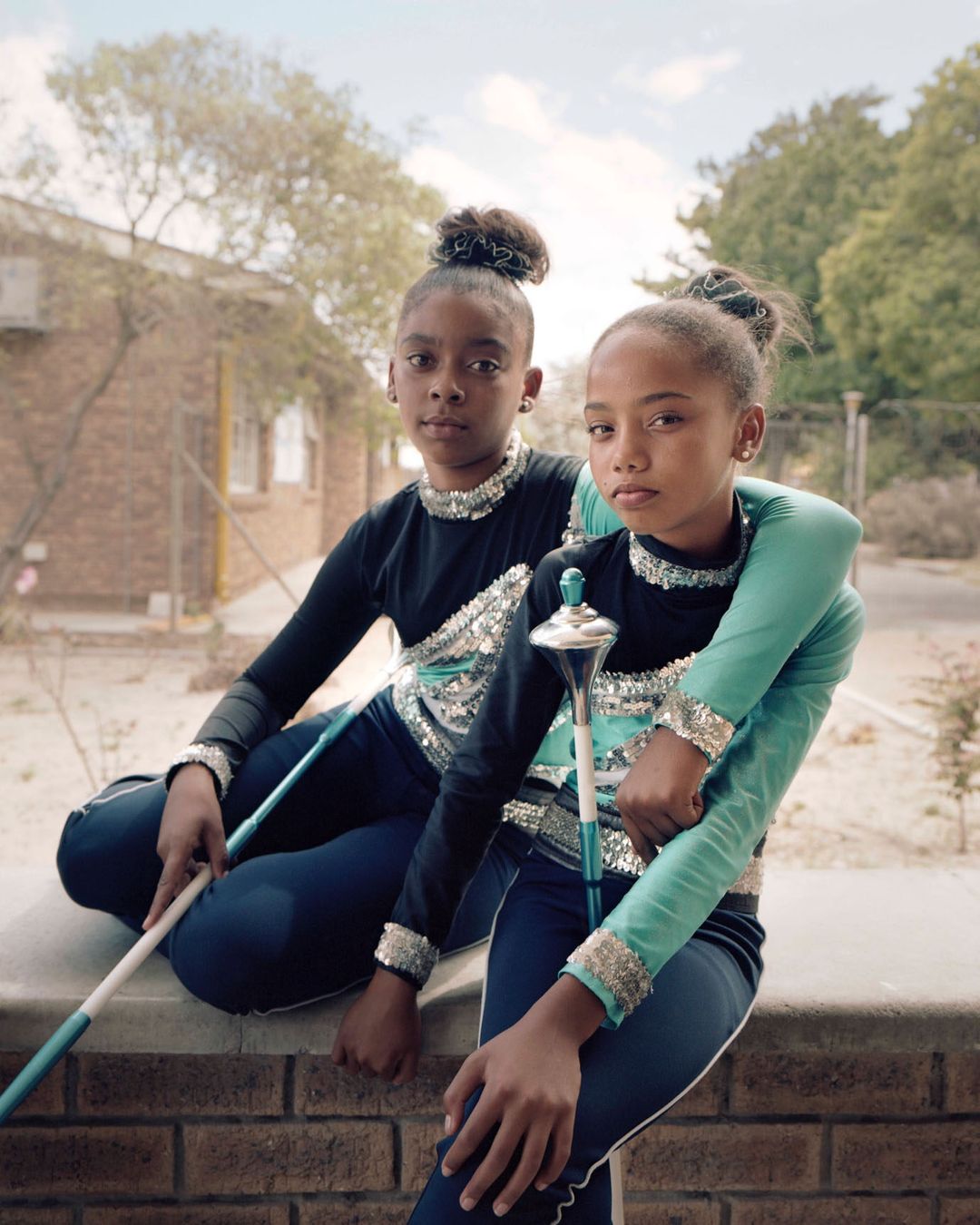 Two young girls in matching turquoise and black uniforms pose while sitting on a brick wall in a medium shot.