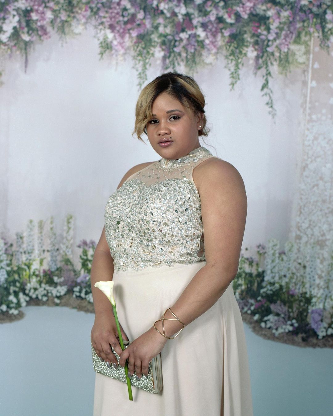Elegant woman in a shimmering gown holds a calla lily and clutch against a floral backdrop in a formal portrait with soft lighting.