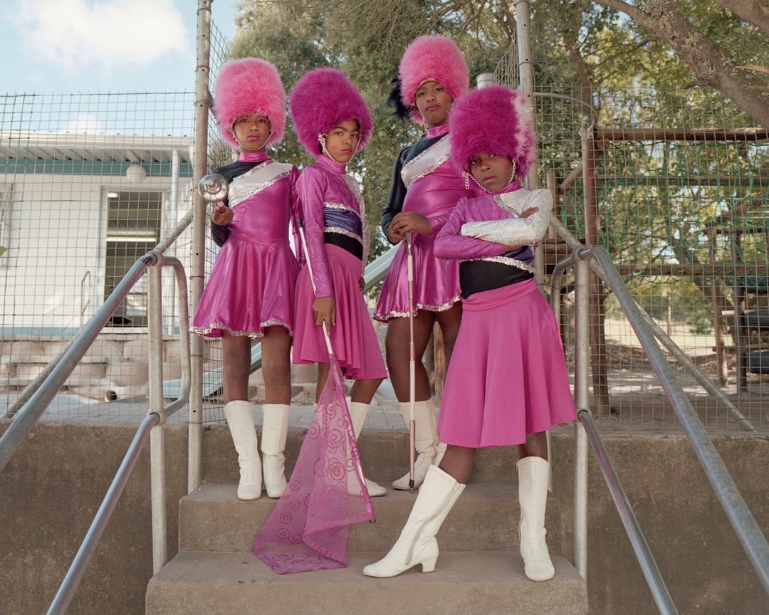 Four children in pink band uniforms stand on stairs in an outdoor setting, two holding batons, showcasing a unique group performance.