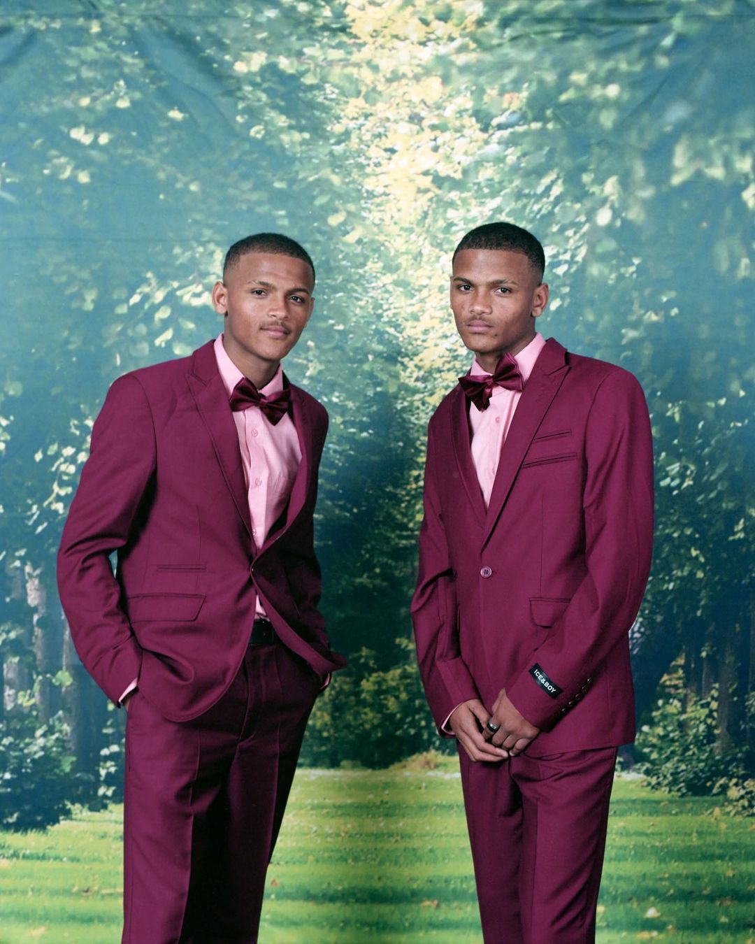 Two men in burgundy suits with bowties pose in front of a background that resembles a path through trees and greenery.