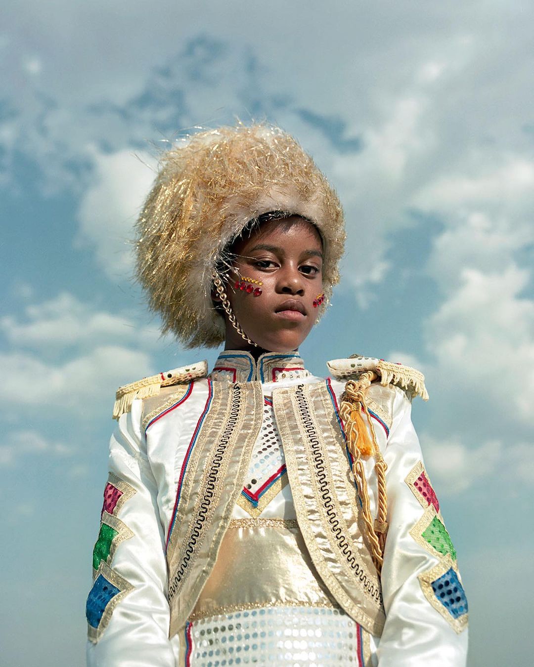 Person wearing a decorative hat and a white and gold jacket with multicolored accents against a partly cloudy sky. Portrait shot.